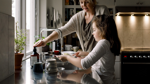 En mamma och ett barn diskar tillsammans vid en köksbänk med rinnande vatten.