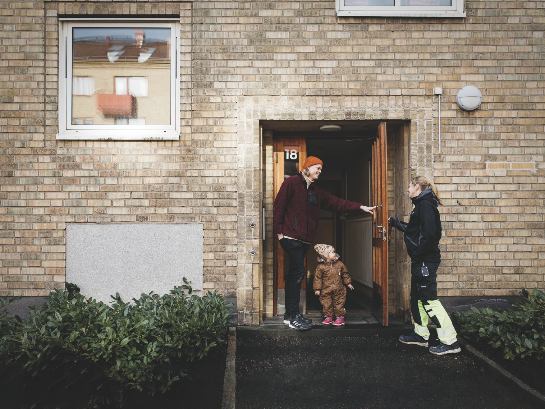 En ung pappa på väg ut från en port tillsammans med sin lilla dotter håller upp dörren för en fastighetsarbetare på Mölndalsbostäder.