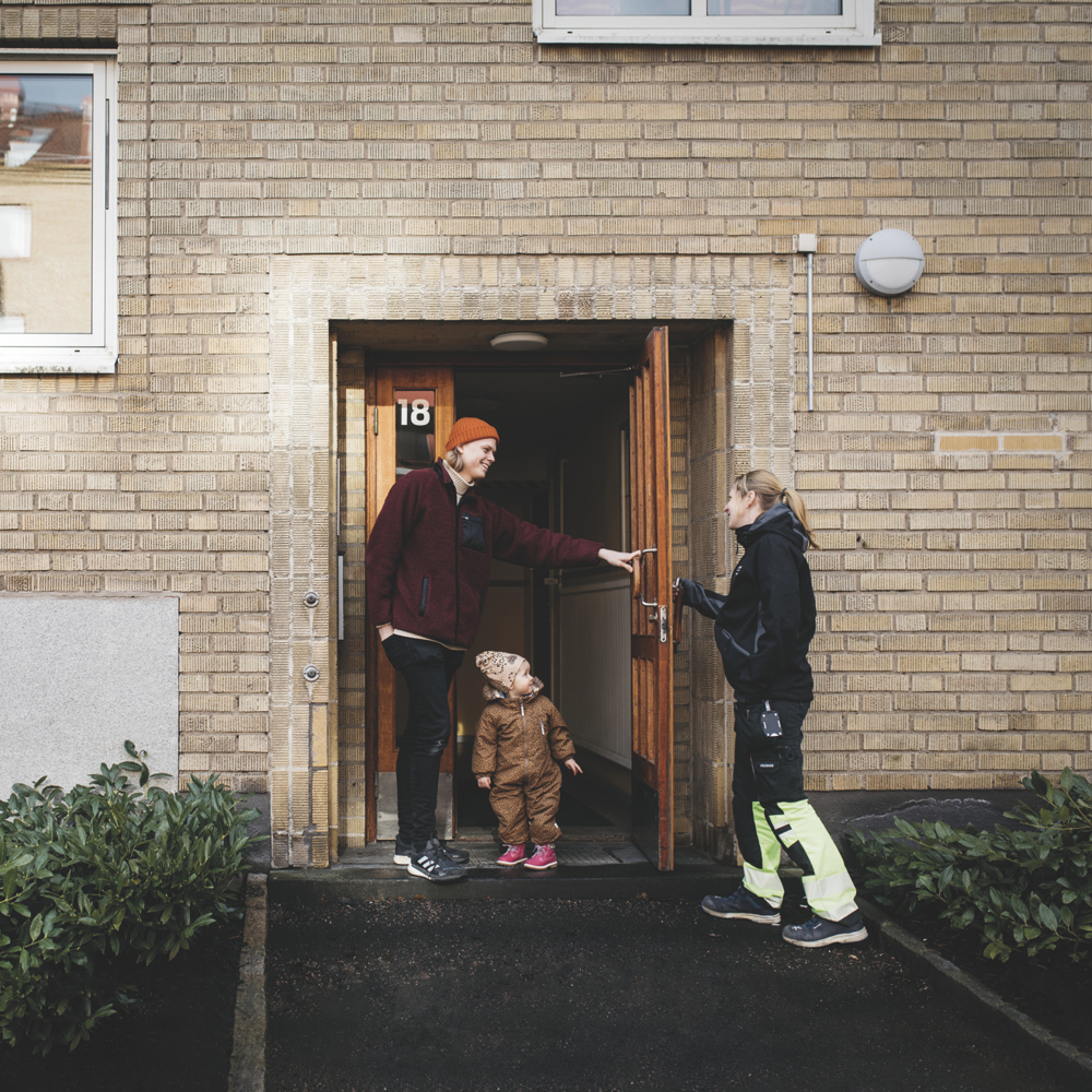 En ung pappa på väg ut från en port tillsammans med sin lilla dotter håller upp dörren för en fastighetsarbetare på Mölndalsbostäder.