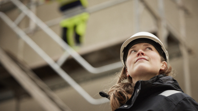Kvinna med bygghjälm med en byggställning i bakgrunden. 