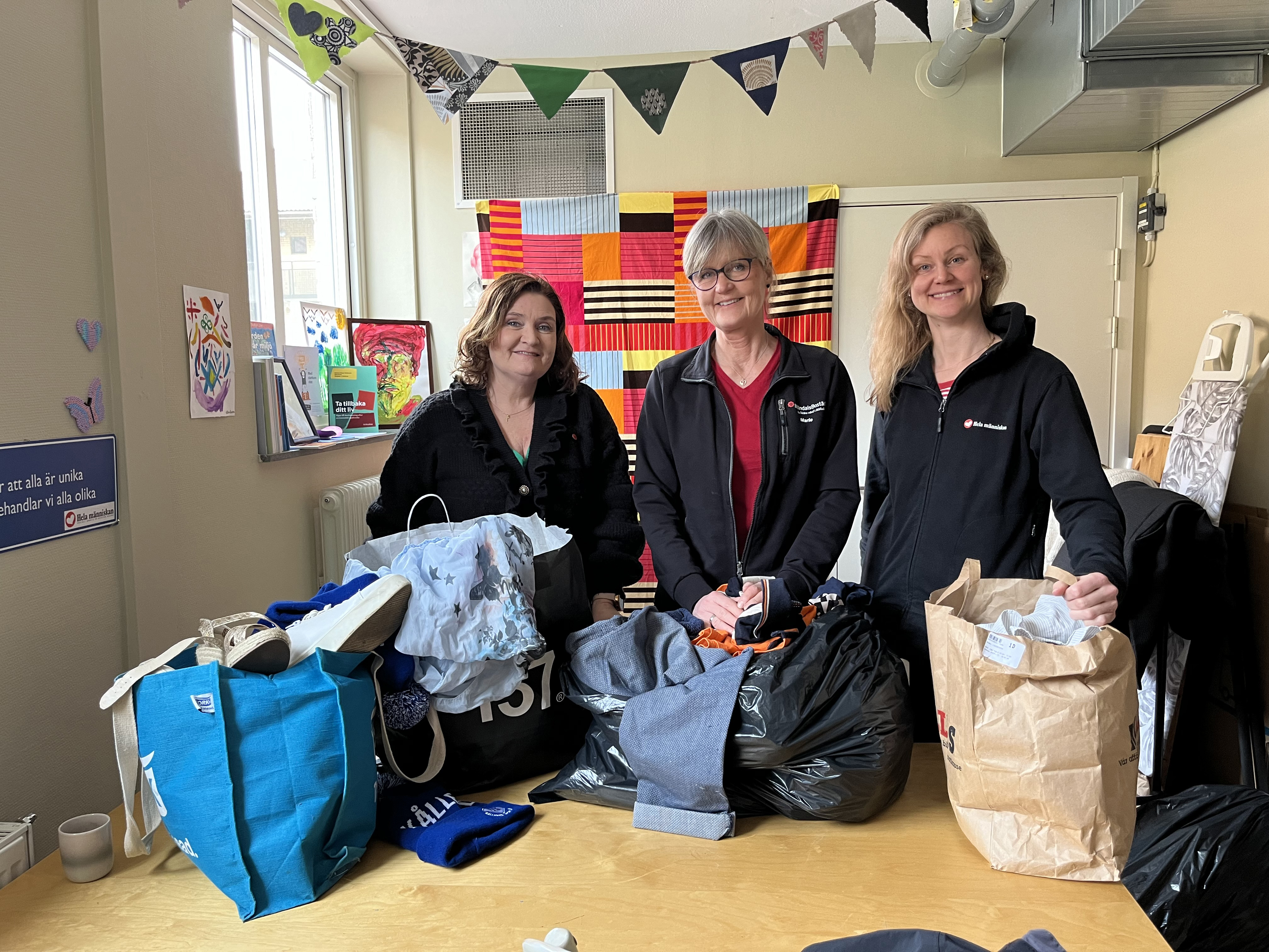 Tre kvinnor står leende tillsammans vid ett bord med kassar som innehåller skänkta kläder. 