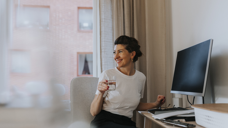 Mörkhårig leende kvinna iklädd vit t-shirt med uppsatt hår sitter bortvänd från en dator i ett hem med ett glas i handen. 