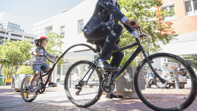 En pappa cyklar med med sitt barn på en cykel anpassad för två i stadsmiljö