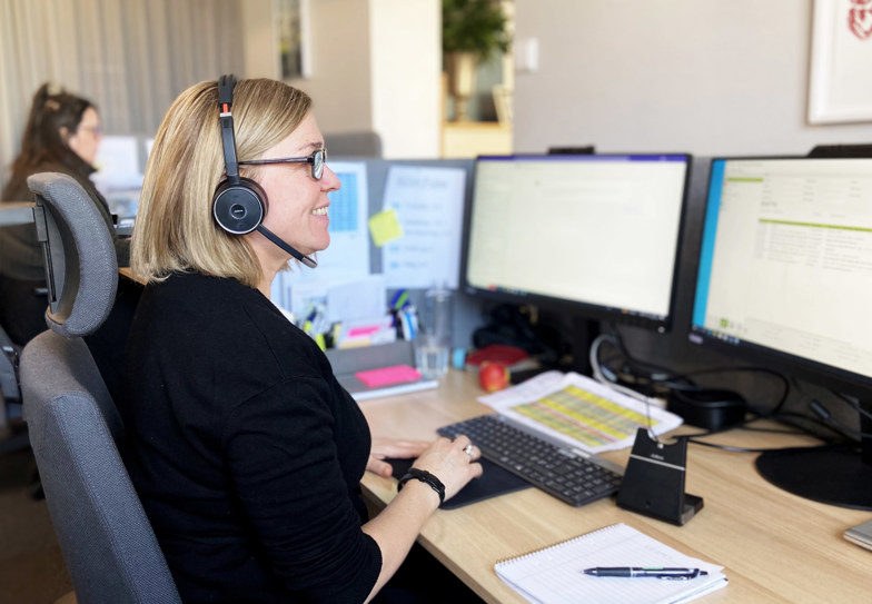 Mellanblond kvinna med telefonheadset ler och tittar på en datorskärm på ett kontor i grått.