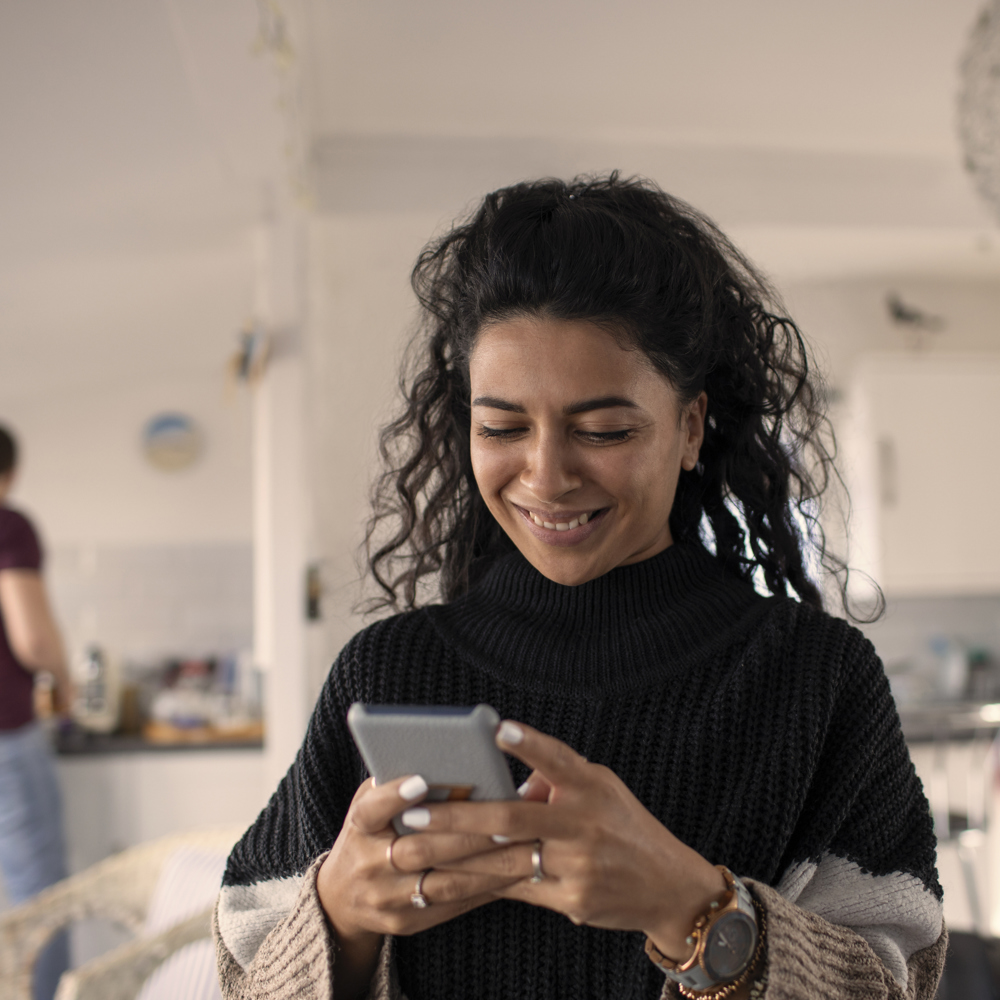 Leende kvinna sitter i sitt vardagsrum och gör sina boendeärenden via sin smartphone. 