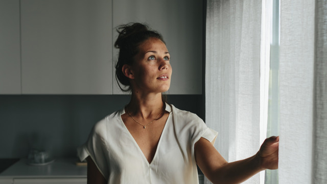 En ung mörkhårig kvinna står vid ett fönster i ett vitt hem och drar för en gardin. 