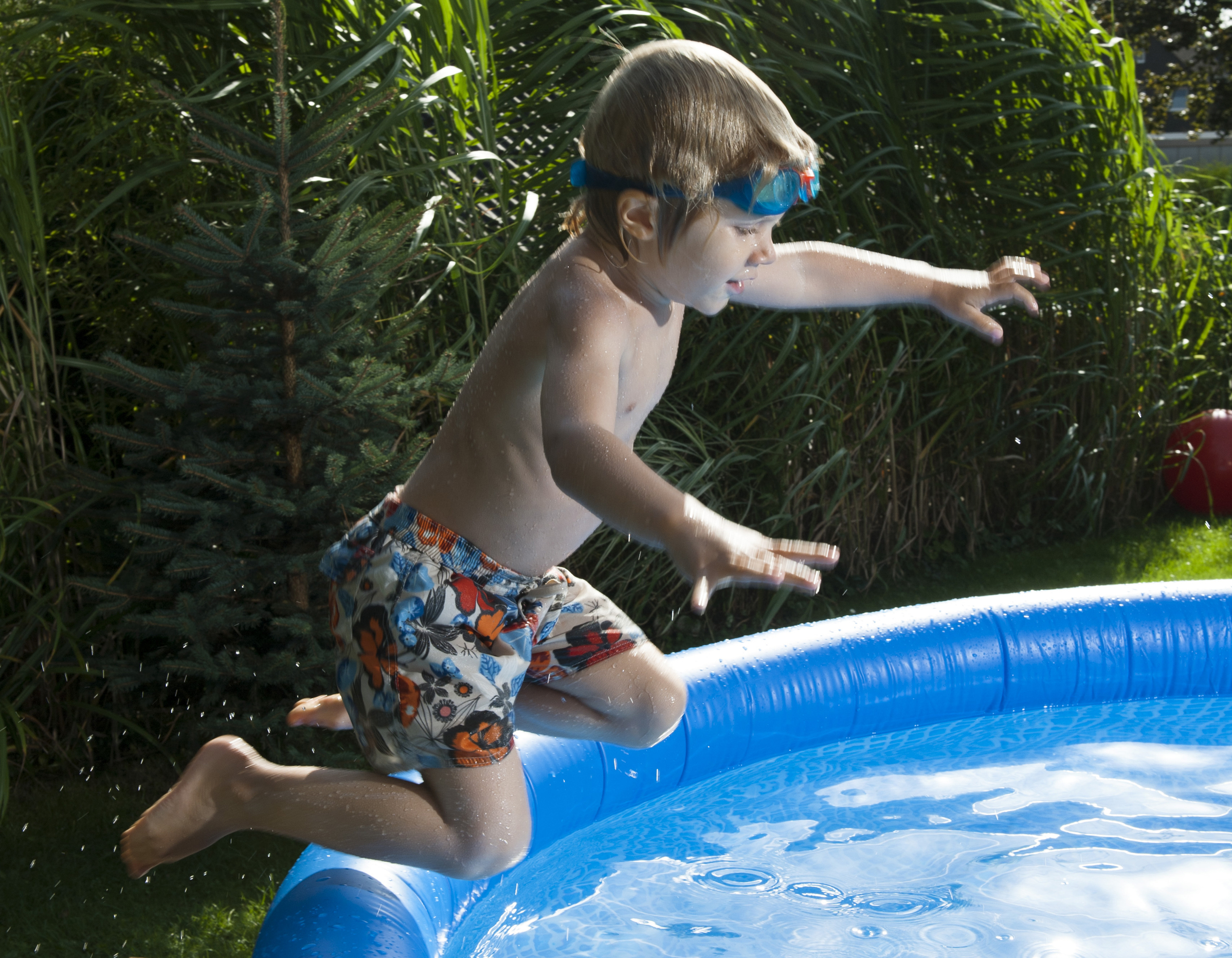 Litet barn med cyklop i pannan och badbyxor hoppar ner i en liten barnbassäng i trädgården. 