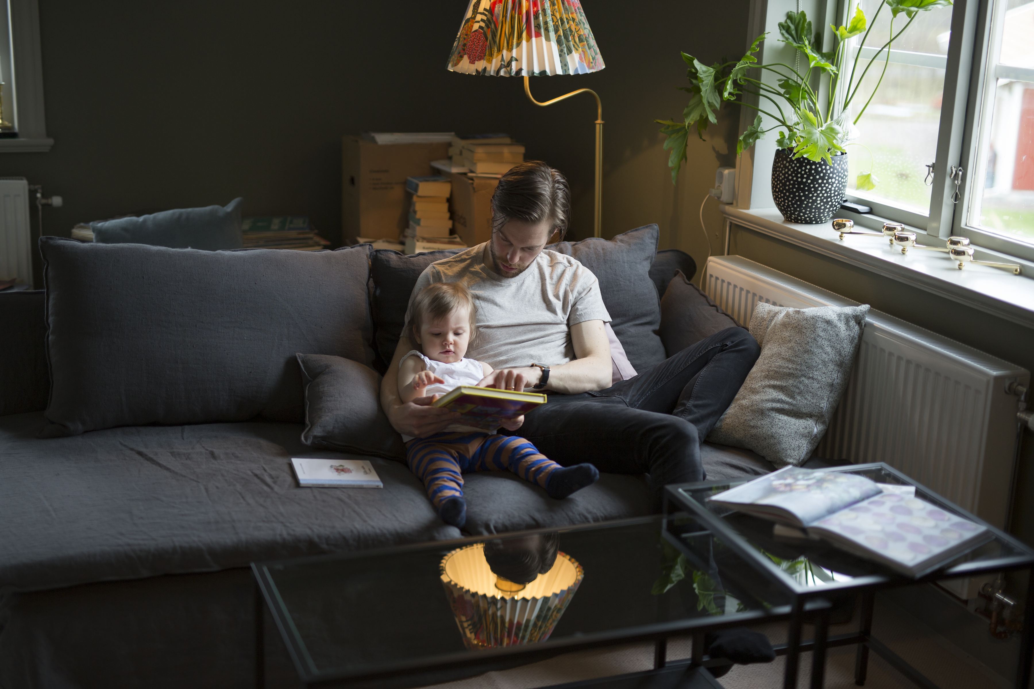 En pappa sitter och läser för sitt lilla barn i skenet av en lampa i en soffa. 
